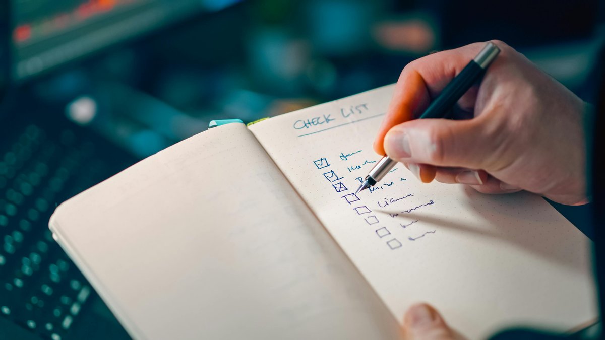 A hand writing a checklist in a notebook beside a keyboard — evaluating readiness item by item
