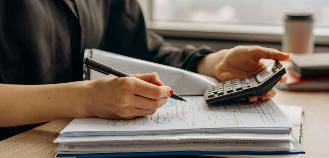 A person reviewing a stack of paper documents with a pen and calculator